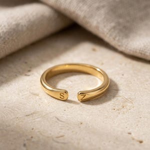 Puede incluir: Un anillo abierto de tono dorado con un acabado liso y pulido. Los dos extremos de la banda están ligeramente aplanados y cada uno muestra una letra grabada, con la letra S en un lado y la letra Z en el otro lado de la abertura.
