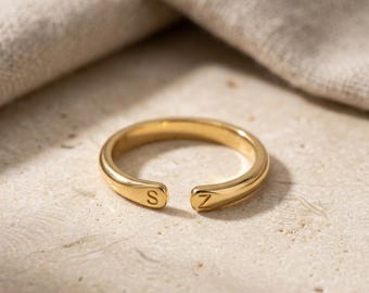 May include: A gold-toned open ring featuring a smooth, polished finish. The two ends of the band are slightly flattened and each displays a single engraved letter, with the letter S on one side and the letter Z on the other side of the opening.