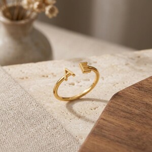 May include: A gold-toned open ring featuring two capital letters, A and E, positioned at the ends of the band. The ring has a smooth, polished finish and a simple, thin circular band design that remains open at the top between the two letters.