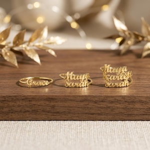 May include: Three gold-toned rings featuring cursive script lettering. The first ring displays the name Grace, the second ring features the names Maya and Sarah stacked, and the third ring displays the names Maya, Scarlett, and Sarah stacked.