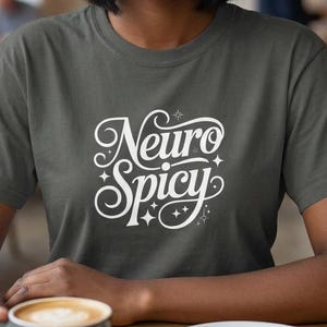 Könnte beinhalten: Eine Frau trägt ein dunkelgraues T-Shirt mit dem weißen Schriftzug "Neuro Spicy". Das Shirt hat einen Rundhalsausschnitt und kurze Ärmel. Die Frau lächelt und hält eine Kaffeetasse in einem Café.