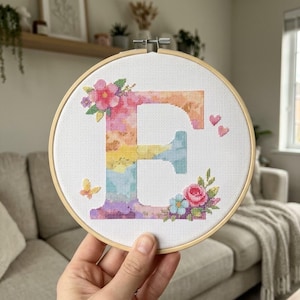 Könnte beinhalten: Ein runder Holzstickrahmen mit weißem Aida-Stoff, der ein Kreuzstichmotiv des Buchstabens E zeigt. Der Buchstabe ist in einem aquarellartigen Regenbogenverlauf gestaltet und mit rosa und blauen Blumen, grünen Blättern sowie einem kleinen Schmetterling verziert.