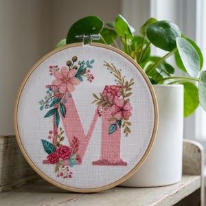 Puede incluir: Un bastidor de bordado circular de madera que contiene tela Aida blanca con la letra M bordada en punto de cruz en color rosa claro. La letra está decorada con arreglos florales bordados con flores y hojas rosas, rojas, verde azulado y verdes.