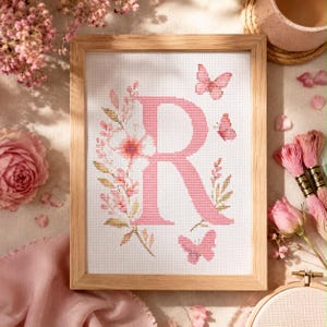 May include: A cross-stitch embroidery piece framed in light wood, featuring a large pink letter R decorated with delicate floral vines and three small pink butterflies on a white fabric background, creating a soft and detailed needlework design.