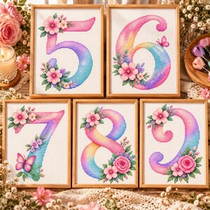 以下が含まれることがあります： 数字の5、6、7、8、9をあしらった5つのクロスステッチ刺繍アート。各数字は鮮やかなレインボーグラデーションで表現され、白い布地に詳細なピンクの花のモチーフと小さな蝶が刺繍されています。