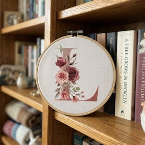 May include: A circular wooden embroidery hoop containing white Aida fabric with a cross-stitched letter L in a muted rose color. The letter is decorated with a cluster of embroidered roses and leaves in shades of deep burgundy and soft pink.