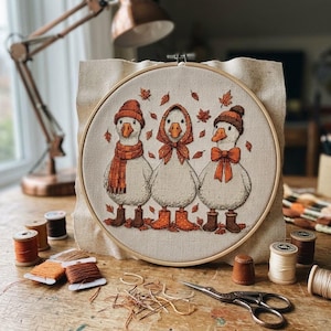 May include: A circular wooden embroidery hoop containing a cross-stitch design of three white geese wearing orange and brown autumn-themed hats, scarves, and boots, surrounded by scattered orange maple leaves on a light beige fabric background.
