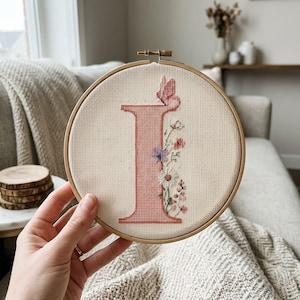 May include: A circular wooden embroidery hoop containing a cross-stitch design on off-white fabric. The design features a large, solid pink letter I decorated with a small pink butterfly at the top and a cluster of colorful wildflowers along the right side.