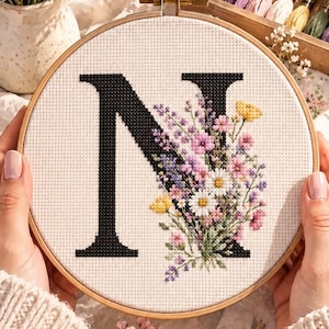 May include: A circular wooden embroidery hoop containing white Aida fabric with a cross-stitched black letter N. The letter is decorated with a colorful floral arrangement featuring pink, purple, white, and yellow flowers with green leaves.