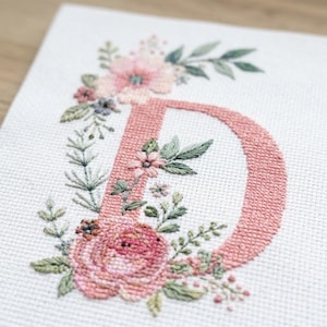 May include: A cross-stitch embroidery piece on white Aida fabric featuring the letter D in a dusty rose colour. The letter is decorated with a floral arrangement of pink and peach flowers, green leaves, and small sprigs in various shades of green.