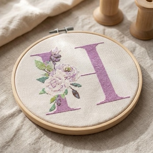 Puede incluir: Un bastidor de bordado circular de madera que contiene una tela de color blanco roto con un diseño de punto de cruz. El diseño presenta una letra H grande de color morado decorada con flores moradas y blancas y hojas verdes.