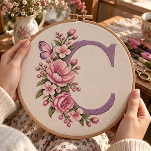 May include: A circular wooden embroidery hoop containing a piece of white fabric with a cross-stitch design. The design features a large purple letter C decorated with pink flowers, green leaves, and a small purple butterfly on the left side.