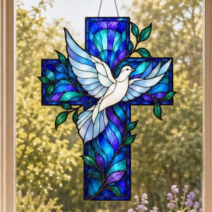 Op de afbeelding: Een glas-in-lood zonnevanger in de vorm van een kruis met een witte duif in vlucht in het midden. Het kruis is samengesteld uit diverse tinten blauw en paars glas met groene bladaccenten. Het hangt aan een dunne metalen ketting voor een raam.