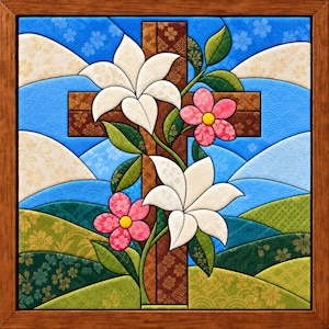 May include: A square framed fabric art piece featuring a brown patterned cross as the central focus. White lilies and pink flowers with yellow centres are intertwined with green leaves across the cross, set against a blue and green landscape.