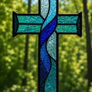 Può includere: Una croce in vetro colorato con un motivo centrale a onde verticali in blu scuro e azzurro, fiancheggiata da bracci orizzontali in vetro testurizzato color ottanio. L'intero pezzo è tenuto insieme da linee di piombo in metallo scuro.