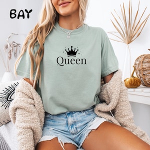 Peut inclure: Un t-shirt à manches courtes vert sauge avec un motif noir représentant une couronne au-dessus du mot Queen écrit dans une police serif. Le t-shirt a un col rond et une coupe décontractée, porté avec un short en jean bleu clair.