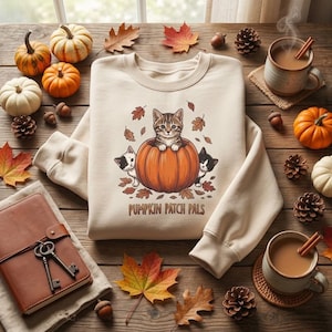 May include: A beige long-sleeve sweatshirt featuring a graphic of three kittens and a large orange pumpkin surrounded by autumn leaves. The text PUMPKIN PATCH PALS is printed below the illustration in a brown, rounded, sans-serif font.