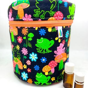 Puede incluir: Un contenedor cilíndrico de tela negra con cierre de cremallera y un estampado colorido de ranas, setas y flores. Dos pequeños frascos de vidrio ámbar con tapas blancas están delante del contenedor.