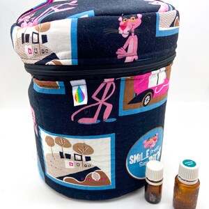 Peut inclure: Un étui cylindrique en tissu noir pour huiles essentielles, orné d'un motif répété de la Panthère Rose dans diverses poses et scènes. L'étui est doté d'une fermeture éclair, d'une poignée et de deux petits flacons d'huiles essentielles.