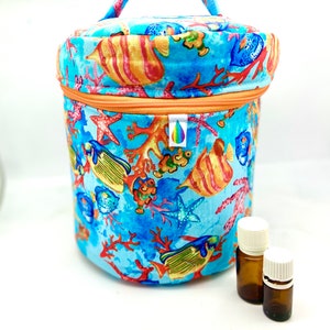 Puede incluir: Un estuche cilíndrico de tela para aceites esenciales con un diseño acuático colorido. El estuche presenta un fondo azul con corales, estrellas de mar y peces. Tiene cierre de cremallera y un asa. Dos pequeñas botellas marrones con tapas blancas están al lado.