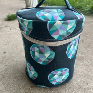 Peut inclure: Sac de rangement cylindrique avec une poignée, orné d'un motif répété de formes géométriques de pierres précieuses turquoise, violettes et crème sur fond noir. Il possède une fermeture éclair beige et une poignée assortie.