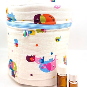 Peut inclure: Un sac cylindrique en tissu crème pour huiles essentielles, avec une fermeture éclair bleue et une poignée blanche. Le sac est décoré d'illustrations d'éléphants colorés. Deux petites bouteilles en verre ambré avec des bouchons blancs sont devant le sac.