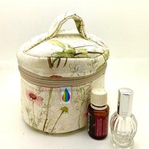 Puede incluir: Un neceser beige con estampado floral, asa, cremallera y logotipo arcoíris. Junto a la bolsa hay dos botellas: una marrón con tapón blanco y otra transparente y decorativa.