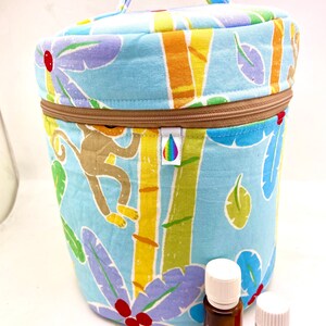 Peut inclure: Une trousse de transport en tissu bleu et jaune avec une fermeture à glissière et une poignée. La trousse est imprimée d'un motif de singe et de feuilles tropicales. Deux petits flacons en verre ambré avec des bouchons blancs sont devant la trousse.