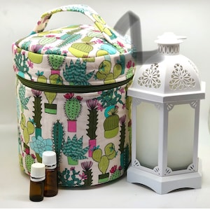 May include: A round, fabric essential oil carrying case with a cactus print in green, pink, and blue on a cream background. A white lantern and three small amber glass bottles are next to the case. The case has a green zipper and a handle.