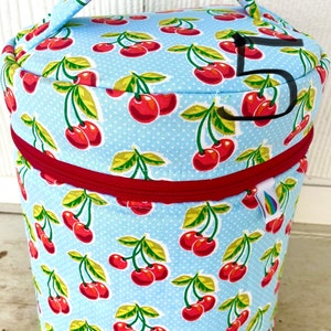 Peut inclure: Un récipient cylindrique avec une fermeture éclair rouge et une poignée. Le récipient est recouvert d'un tissu bleu clair avec un motif de cerises rouges et de feuilles vertes. Des pois blancs sont dispersés sur le tissu.