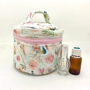 Puede incluir: Un neceser cilíndrico con estampado floral, cremallera y asa rosas. La bolsa presenta un patrón de flores y plumas. Junto a la bolsa hay dos botellas, una transparente y otra marrón.