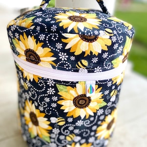 Puede incluir: Un contenedor de almacenamiento de tela negro con una cremallera blanca y un asa. El contenedor está decorado con un patrón de girasoles y abejas.