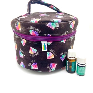 May include: A round black fabric storage case with a purple zip and a top handle. The fabric features a repeating pattern of colourful diamond illustrations and the text "SHINE LIKE A". A small white tag with a rainbow droplet icon is attached.