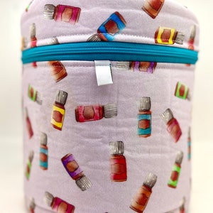 Peut inclure: Un étui cylindrique lavande pour huiles essentielles avec une fermeture éclair et une poignée turquoise. L'étui est recouvert d'un motif répété de flacons d'huiles essentielles de différentes couleurs, dont le marron, le rouge, le jaune et le bleu.