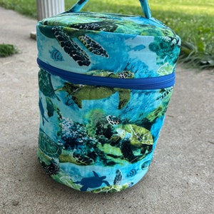 Peut inclure: Un sac cylindrique en tissu bleu et vert avec une fermeture à glissière et une poignée. Le sac présente un imprimé de tortues marines et de poissons.