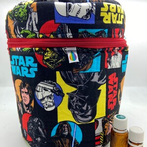 Peut inclure: Un contenant de rangement en tissu noir avec une fermeture éclair rouge et un imprimé Star Wars mettant en scène des personnages comme Dark Vador, Yoda et Luke Skywalker. Deux petits flacons en verre brun avec des bouchons blancs sont au premier plan.