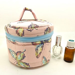 Puede incluir: Un neceser redondo de tela rosa con cremallera azul y asa. La bolsa está decorada con grandes estampados de mariposas en tonos amarillos, azules y morados. Dos frascos de vidrio están junto a la bolsa.