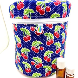 May include: A cylindrical, fabric essential oil carrying case with a cherry pattern on a navy blue background. The case has a white zipper, a handle, and a small tag. Two small brown glass bottles with white lids are next to the case.