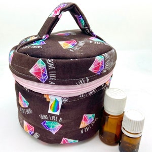 Könnte beinhalten: Eine braune runde Reißverschlusstasche mit einem Griff. Die Tasche hat einen rosa Reißverschluss und ist mit einem sich wiederholenden Muster aus regenbogenfarbenen Diamanten bedeckt, auf denen der Text "Shine Like A" gedruckt ist. Zwei kleine Glasflaschen stehen vor der Tasche.