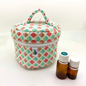 Puede incluir: Un estuche redondo para aceites esenciales con asa trenzada. El estuche presenta un diseño geométrico en coral, verde menta y dorado sobre fondo blanco. Dos pequeñas botellas de vidrio marrón con tapas blancas.
