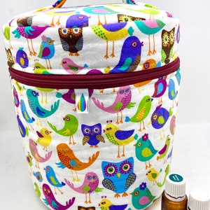Puede incluir: Una bolsa redonda de tela blanca con cierre de cremallera y asa. La bolsa está decorada con un patrón colorido de pájaros y búhos. Dos pequeñas botellas de vidrio marrón con tapas blancas están delante de la bolsa.