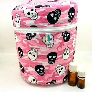 Peut inclure: Un sac rond rose à motif camouflage avec une fermeture éclair blanche et une poignée. Le sac est décoré de motifs de crânes noirs et blancs. Deux petites bouteilles en verre brun avec des bouchons blancs sont à côté du sac.