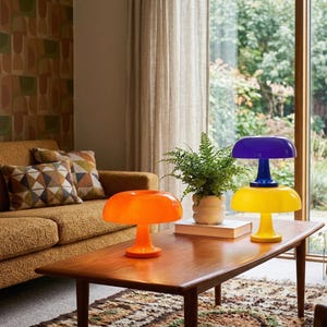 May include: A living room scene features a brown sofa with patterned pillows and a wooden coffee table. Two mushroom-shaped lamps in orange and yellow with blue accents sit on the table. A fern is in a white pot.