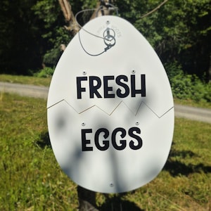 May include: A white, egg-shaped metal sign featuring a horizontal jagged crack across the middle. The text "FRESH" is printed in black capital letters above the crack, and "EGGS" is printed in black capital letters below the crack. A metal wire loop is attached to the top for hanging.