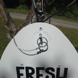 May include: A white, egg-shaped sign featuring the black text "FRESH" and the embossed words "RUNWAY ROOTS" near the top. The sign is suspended by a silver metal wire loop attached to a black carabiner, with a metal pin secured by a ring.