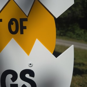 May include: A white and yellow sign shaped like a cracked egg. The white portion features black text with the letters GS visible at the bottom and the letters OF visible in the middle. The yellow center section is smooth and solid in color.
