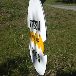 May include: A white, egg-shaped sign with a jagged yellow horizontal crack across the center. The sign features black text that reads FRESH in the top section, OUT OF in the yellow middle section, and EGGS in the bottom section of the egg shape.