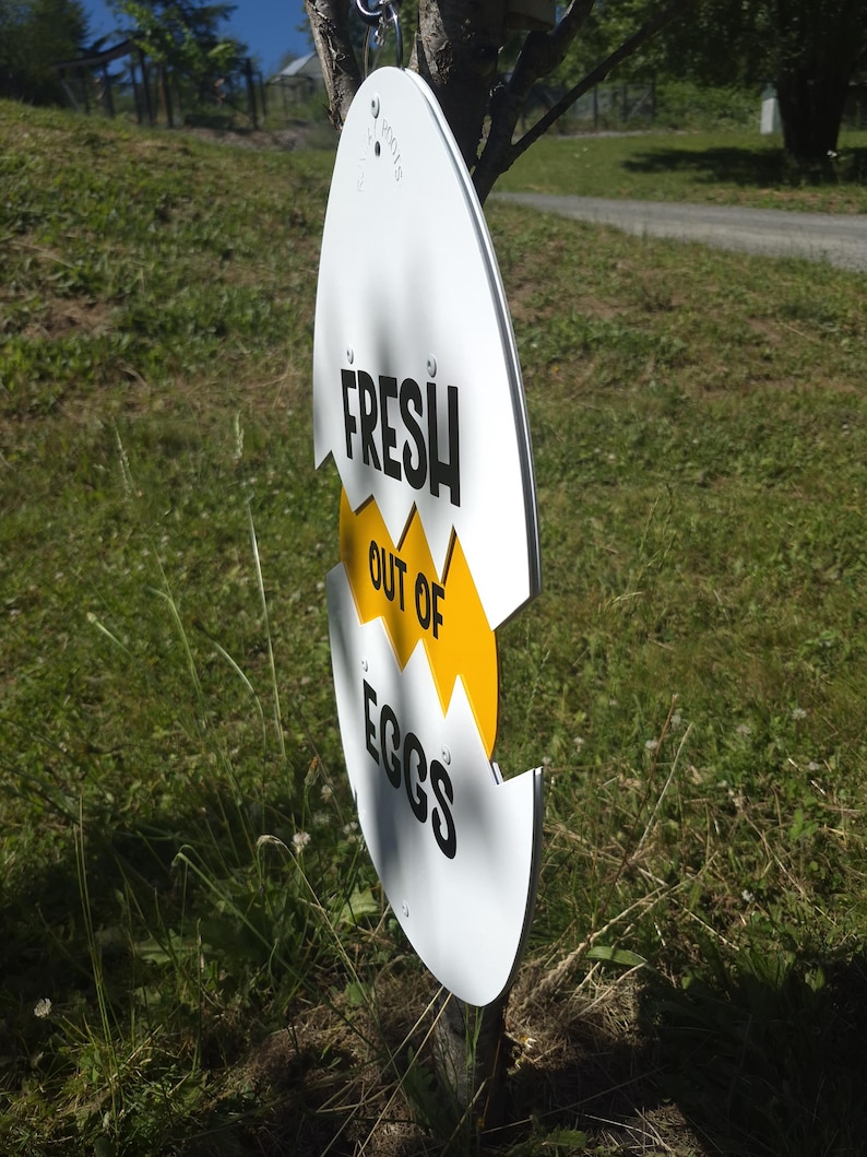May include: A white, egg-shaped sign with a jagged yellow horizontal crack across the center. The sign features black text that reads FRESH in the top section, OUT OF in the yellow middle section, and EGGS in the bottom section of the egg shape.