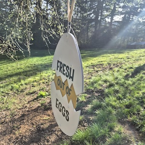 May include: A white, egg-shaped hanging sign with a jagged horizontal crack across the center. The sign features the black text "FRESH OUT OF EGGS" printed on the surface, with a yellow interior visible through the crack, suspended by a tan ribbon.