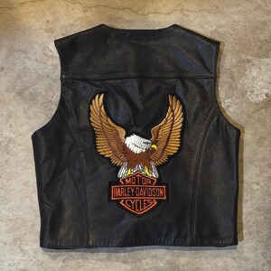 Può includere: Un gilet in pelle nera con una grande toppa ricamata sul retro. La toppa raffigura un'aquila marrone e bianca con le ali spiegate posizionata sopra un logo arancione e nero a forma di scudo con la scritta MOTOR HARLEY-DAVIDSON CYCLES.